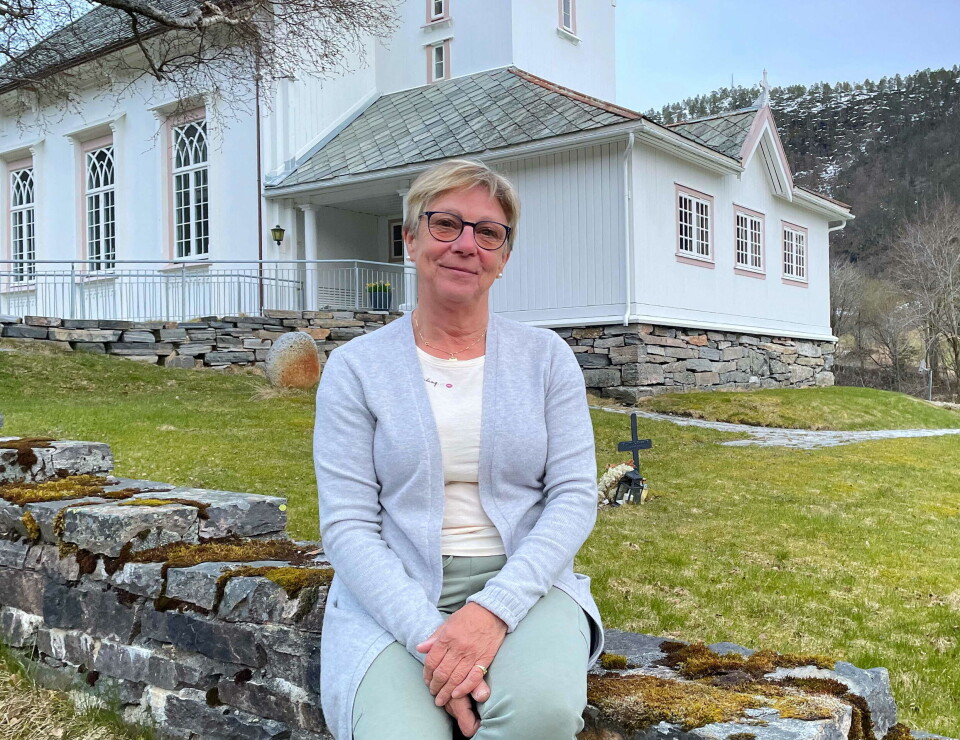 Soknerådsleiar Eva Jorid Svendsen inviterer igjen til gudsteneste og årsfest. Dame sitter på steingjerde foran kirke