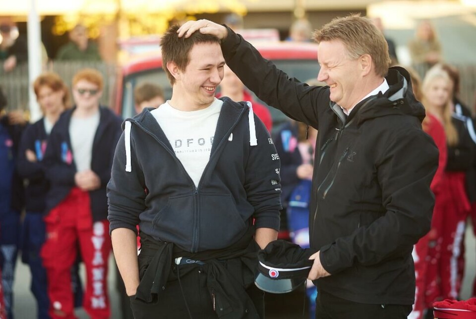 Lattermildt. Mann med hånden på hodet til en russ. Mannen har en russelue i andre hånden. Russ og russebil i bakgrunnen.