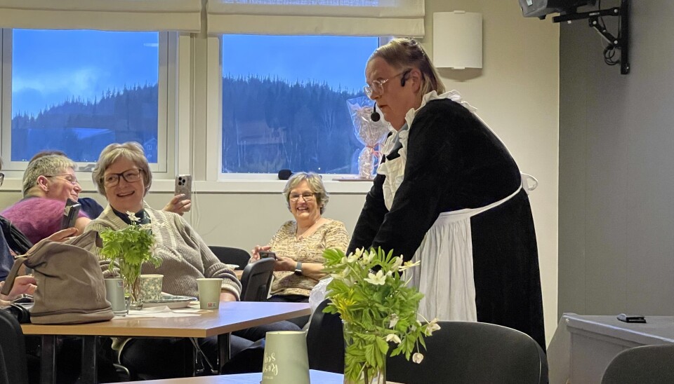 Signe Stunden ankommer salen med fult liv. Hun tar for seg Kirsti Landsem som 'om ikke annet kunne bli frisør, for det krevdes ikke all verden'. I forgrunnen en dame i svart kjole og forkle. Bak sitter noen damer og ler