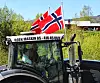 To personer som sitter inne i en traktor og kjører. De har pyntet traktoren med to norske flagg på taket.