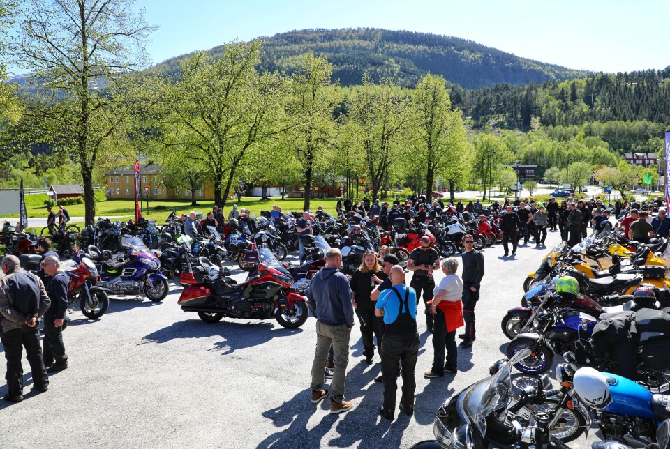 Over 160 motorsyklister besøkte Motorsykkelen dag i Surnadal. Oversiktbilde med full parkeringsplass (av folk og mc) og grønne tre i bakgrunnen.