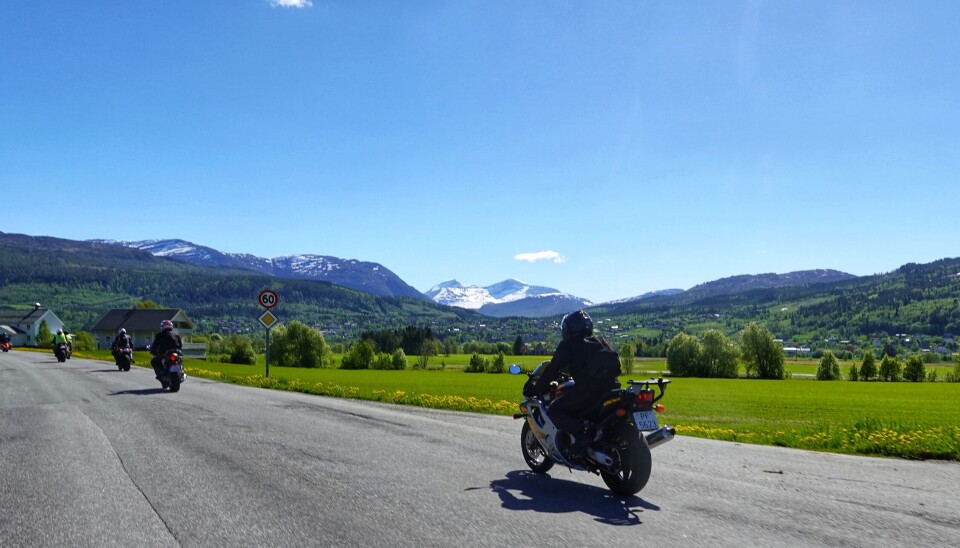 Glimt fra kortesjen. Bilde tatt fra veien, motorsykler bakfra, grønne ekrer ved veien, Surnadal sentrum og fjell i bakgrunnen.