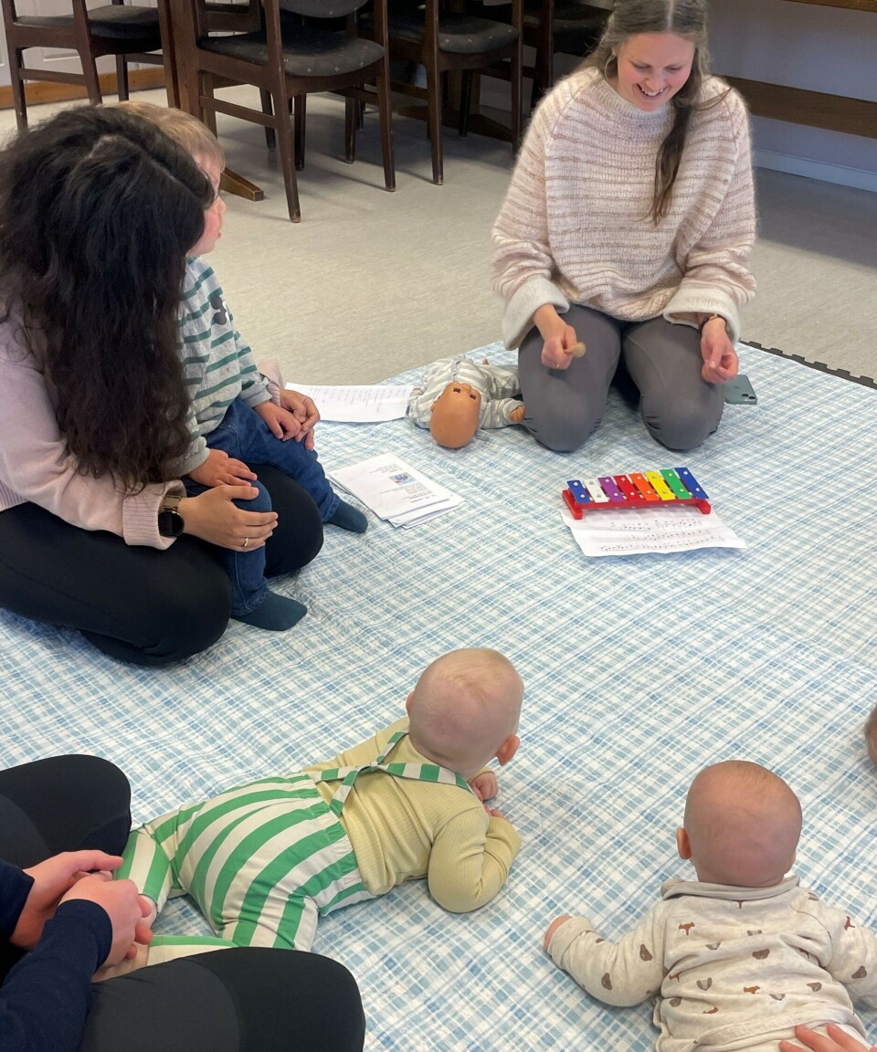 Maria Fiske Eliassen leder babysangen. Voksen og små barn som sitter i ring på et teppe på gulvet.
