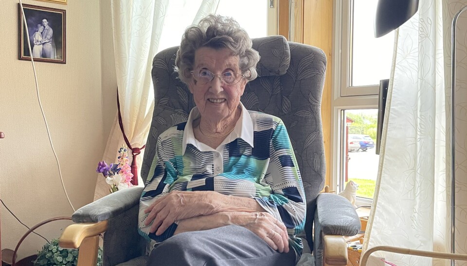 Olga Løfald trives i leiligheten, men savner å være hjemme. En eldre dame sitter i en stol foran ett vindu. har grå bukse og bluse som er rutete