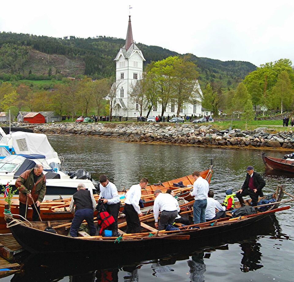Kyrkjebåtroing har prega pinsedagane i Stangvik – som her i 2010. En lang robåt med flere personer om bord, på sjøen, med ei kirke i bakgrunnen.