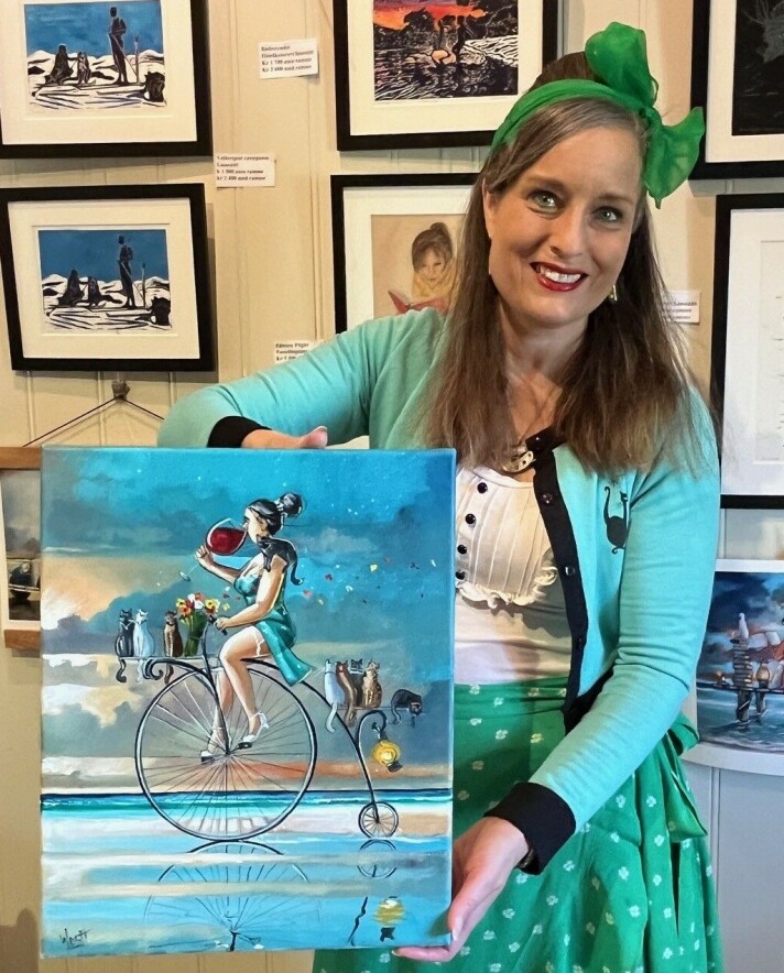 Inga viser et annet av Ronalds malerier, med mange katter på. Ei dame som holder fram et fargerikt maleri.