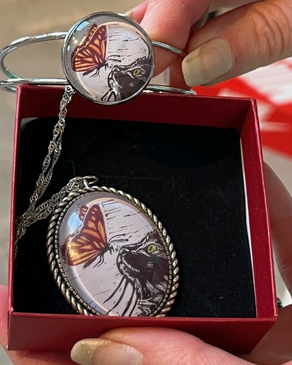 Inga viser et smykke og et armbånd med kattemotiv. Hender som holder opp to smykker.