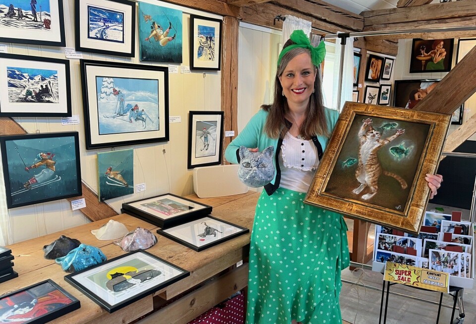 Inga viser et maleri av Signe Førde Bødtker, og et kattehode i keramikk av Ingrid Cimmerbeck. På bordet ligger flere bilder og gjenstander til kattekunst-utstillingen. Ei dame som holder opp et maleri og et kattehode i keramikk.