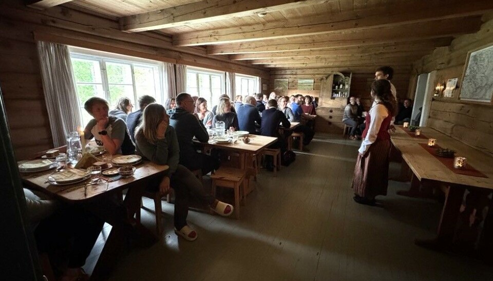 Oda og Amund presenterer dagens middag. Mange personer ved bordene i en spisesal med tømmervegger.
