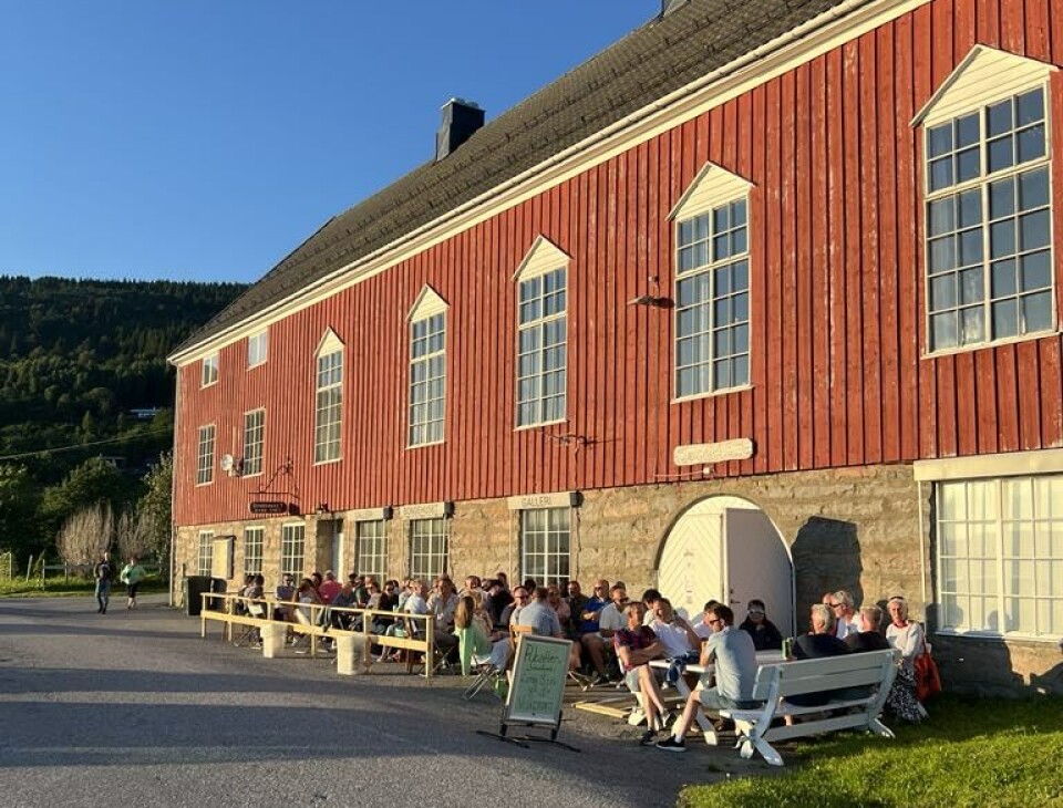 Mange mennesker som sitter ute foran et forsamlingshus på en fin sommerdag/kveld.