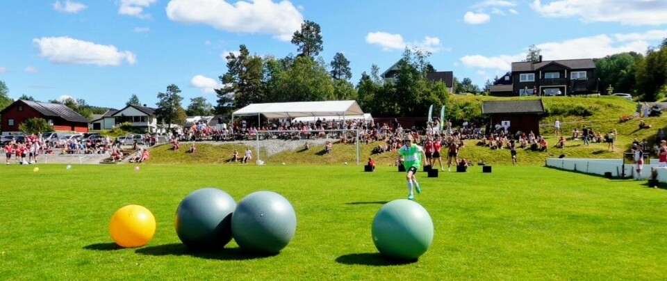 Jente løper mot fire store baller liggende midt på fotballbanen, i bakgrunnen 4 deltagere som ikke er ferdig med posten sin og i skråningen bak sidelinja sitter og står mer enn 100 tilskuere.