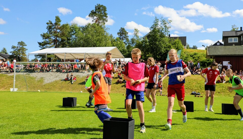 5 barn løper mellom svarte bokser på fotballbanen, rødkledde funksjonærer innimellom. Publikum i bakgrunnen