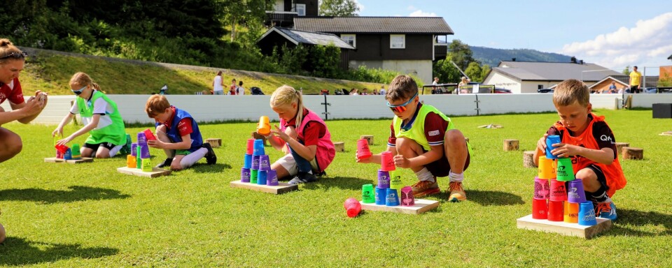 5 barn sitter på huk ved sidelinja av fotballbanen og stabler fargerike kopper.