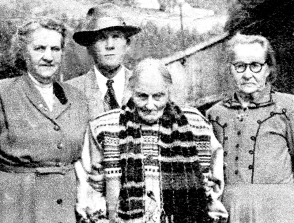 Guro på 105-årsdagen i 1954 – i sjølstrikka kofte og skjerf. Besøkande er Berit og Hilmar Aunli og Dordi Trodal (t.h.). Et gammelt bilde av ei svært gammel dame i strikka kofte. Bak henne står to alvorlige godt voksne damer og en godt voksen mann med hatt.