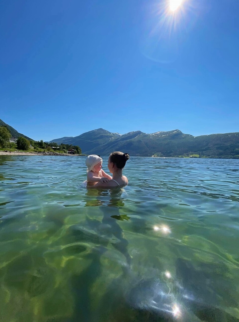 Bildet er tatt på stranda på til Lisbet og Jan Ove Gausen Øigård Norvik, for to år siden. Ei ung dame og et liten barn som bader i klart, gjennomsiktig vann med fjell i bakgrunnen. Sol høgt på skyfri himmel.