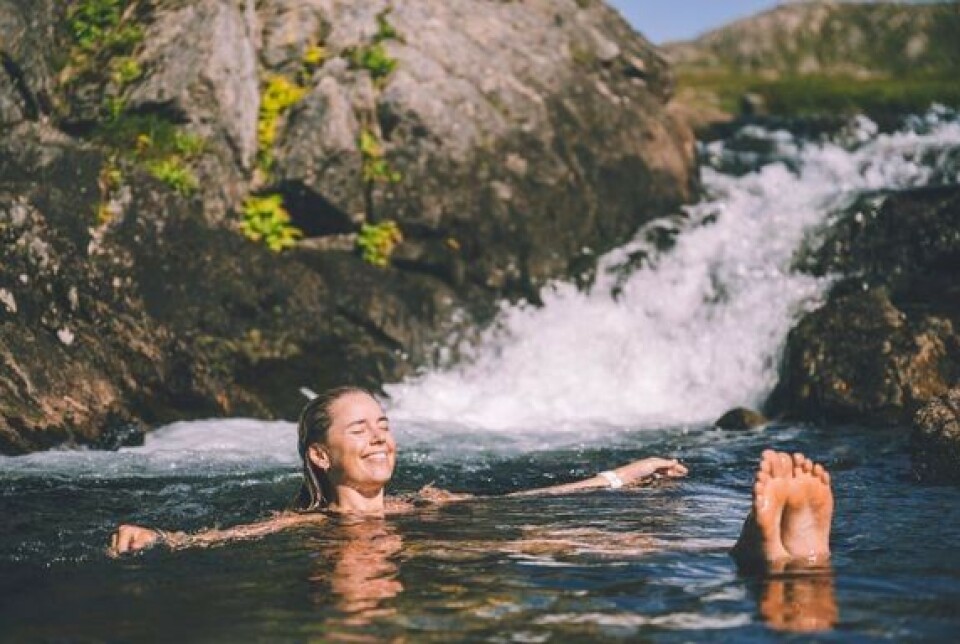 Flere vil ha flere opplevelser i ferien, og gjerne i Norge. Ei dame som bader i ei elv,. Hun ligger smilende på ryggen og flyter i vannet, med hodet og føttene over vannskorpa.