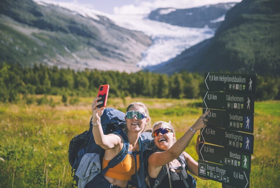 Bilder tatt ved Tåkeheimen To unge damer som tar en selfie ved et skit som viser flere turstier. Man ser fjellet i bakgrunnen.