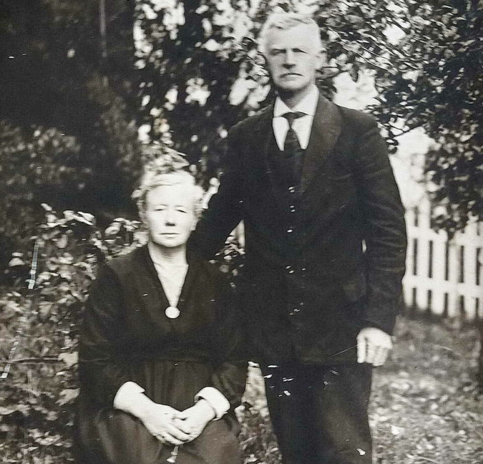 Jordmor Anna Snekvik og mannen Ole John. Gammelt svart/hvitt-bilde av et voksent ektepar, begge med grått hår, i en hage. Hun sitter på en stol, i svart kjole, og med oppsatt hår. Han står ved siden av, i svart dress med slips.