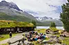 Flere personer som sitter ved rasteplassbord foran et seterhus, med flotte fjell og et vann i bakgrunnen. Sommervær og blå himmel.