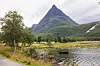Et pyramideformet fjell med et fjellvann i forgrunnen. Overskyet sommervær.