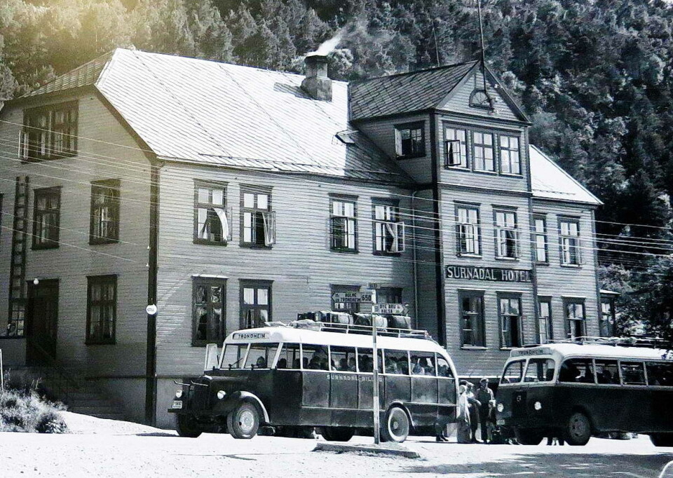 To Trondheims-bussar ved Surnadal Hotell i si tid. Gammelt sort/hvitt bilde av to sorte og hvite busser foran hotell med tre etasjer. Ås i bakgrunnen. Den ene bussen har bagasje på taket.