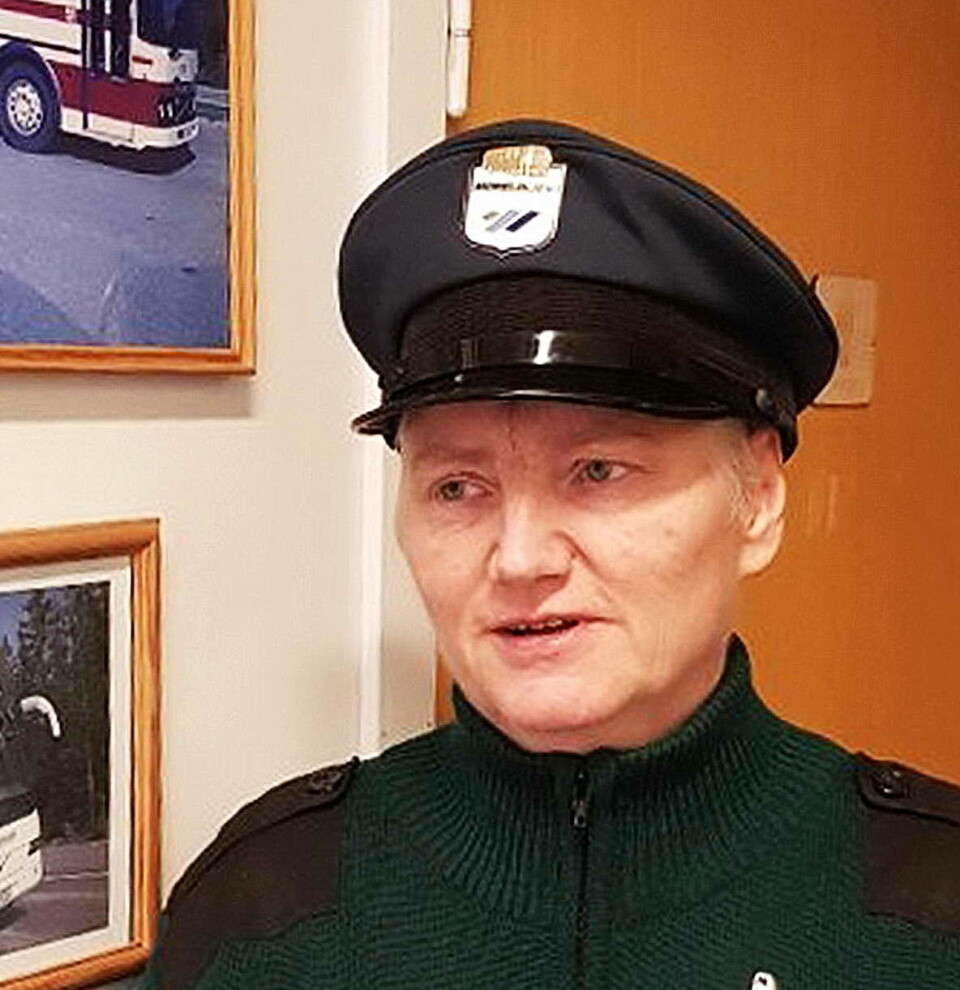 Kanskje har Gerd Anny Gjengstø ei hand på rattet og? Dame i grønn høyhalset jakke med uniformslue. Stpr foran en brun dør og en vegg med to bilder av busser