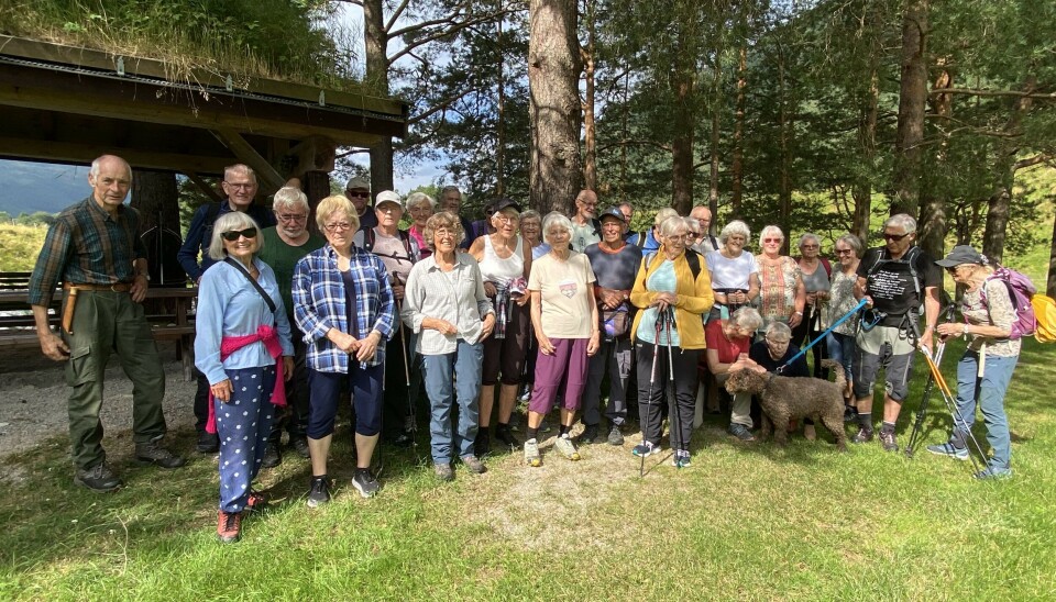Stavgangtur langs Toåa Ei gruppe med ca 20 pensjonister, og en stor hund med krøllete pels, oppstilt for fotografering ved en gapahuk. Sol og sommer.