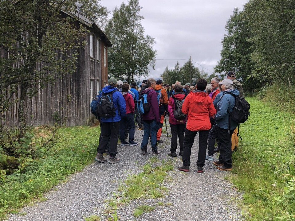 Bergheim/Røsgrinna. Mange turkledde mennesker på en grusvei ved et gammelt hus, i grått og vått sommervær.