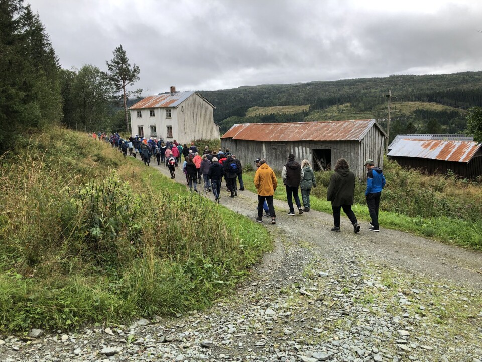 Solberg. Mange turkledde mennesker som går på en grusvei og passerer en gammel gård, i grått og vått sommervær
