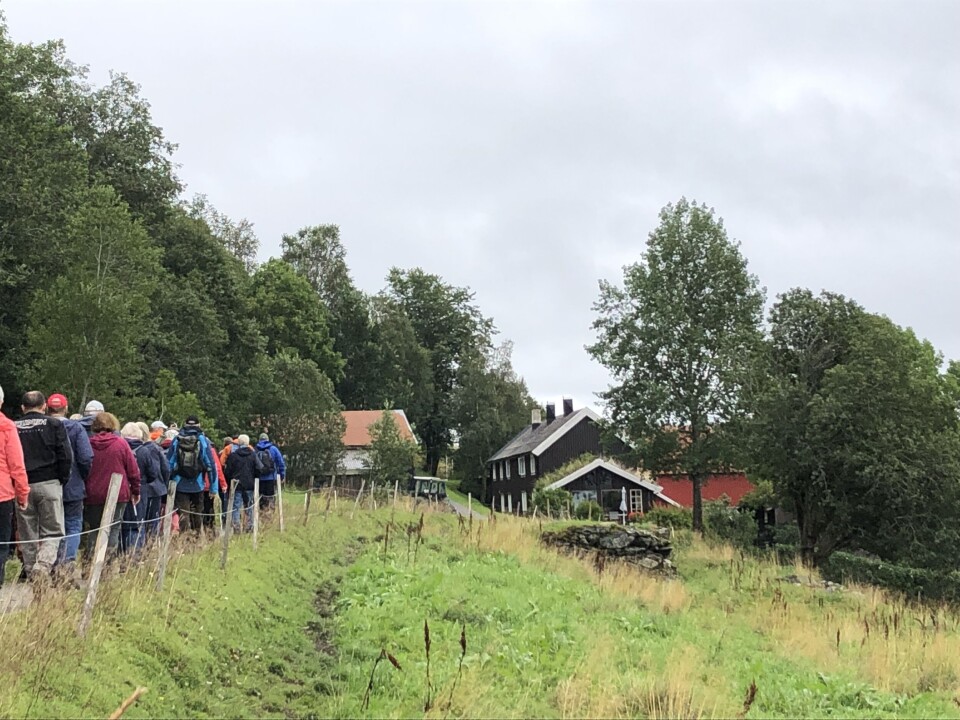 Låvekloppa i Utistu Yttgarden. Ei rekke turkledde mennesker som går på en grusvei, og passerer restene av ei gammel låvebru av stein, grått og vått sommervær.