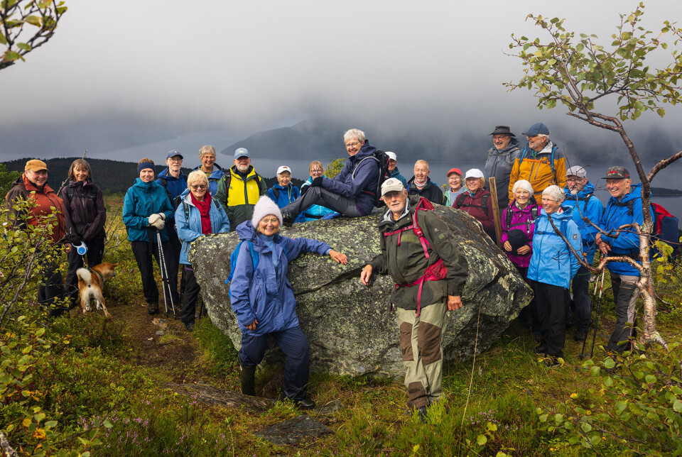 Alle 22 i solglimt ved Middagssteinen. Ei gruppe på 22 turgåere kledd for regnvær, samla rundt en stur stein i marka. I bakgrunnen skimtes fjord og fjell gjennom tåke.