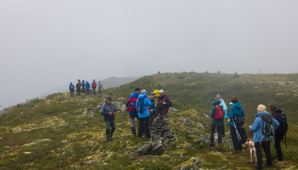 Mot Klokkarhaugen i skoddeeim. Ei gruppe på undt 20 turgåere, kledd for regnvær, og en spisshund, som går i mindre grupper på en tursti på fjellet. Utsikten skjules av tåke.