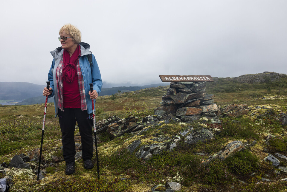 Oppe på Klokkarhaugen. Ei dame med lyst halvlangt hår og briller, kledd for regnvær, står ved en varde på fjellet. Oppå varden ligger et skilt som det står 'klokkarhaugen' på. Tåke i bakgrunnen.