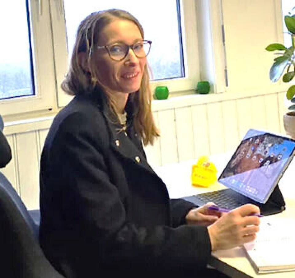 Rannveig Eikrem Dame i blå kåpe, langt lyst hår og briller, sittende på en kontorstol ved en ipad i et rom med vinduer og en grønn plante