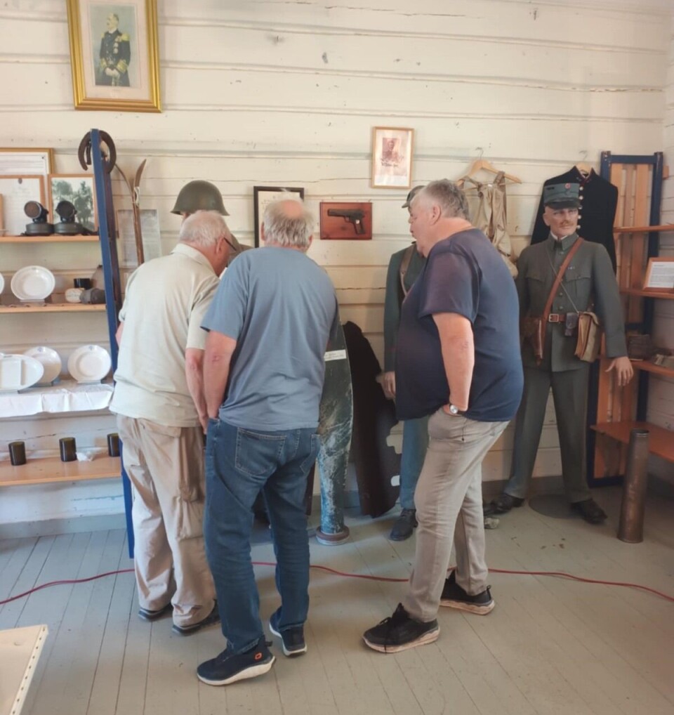 Fullt hus på Åsen Tre menn står med ryggen til og ser på museumsgjenstander fra krigen i et hvitmalt rom. Dukke med uniform til venstre i bildet.