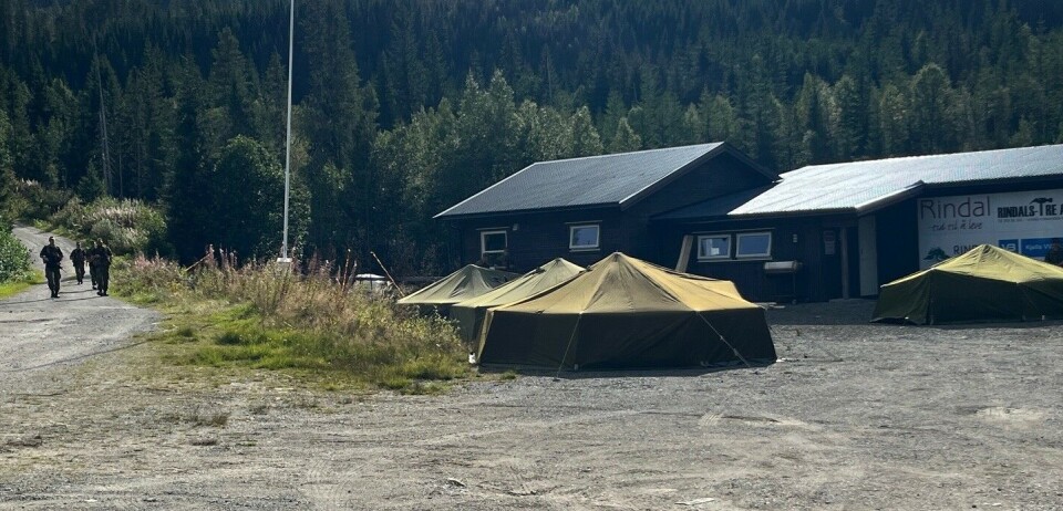 Militærleir ved skytebanen. Fire militære åttemannstelt slått opp foran et hus. Ved siden av kommer noen få soldater gående nedover en vei. Sol og sommer.