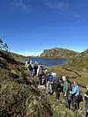 Mennesker i turklær på rekke på sti på fjellet. Vann med bergknauser rundt i bakgrunnen. Blå himmel. Noen få bartrær på venstre side i bildet.