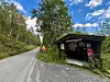 Grusveg som går innover skogen. Skilt med varsel om høy vegkant. Bod på høyre side av vegen med skilt 'Vestre Nordmarksvegen' og informasjon om bompenger.