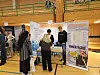 Stand med plakater rundt. To unge menn bak standen, tre jenter foran standen