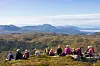 Ei gruppe med flere turkledde personer som sitter samen i en bakke i marka, utsikt over skog og fjell. Sommer og lettskyet himmel.