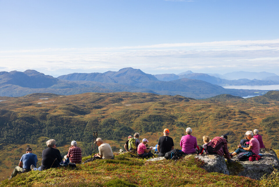 Endeleg oppå fjellet med liten matpause. Ei gruppe med flere turkledde personer som sitter samen i en bakke i marka, utsikt over skog og fjell. Sommer og lettskyet himmel.