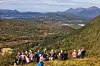 Ei gruppe med flere turkledde personer som sitter samen i en bakke i marka, utsikt over skog og fjell. Sommer og lettskyet himmel.