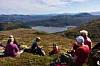 Fem turkledde personer som sitter samen på fjellet, med utsikt over fjellheimen, og et vatn. Sommer og lettskyet himmel.