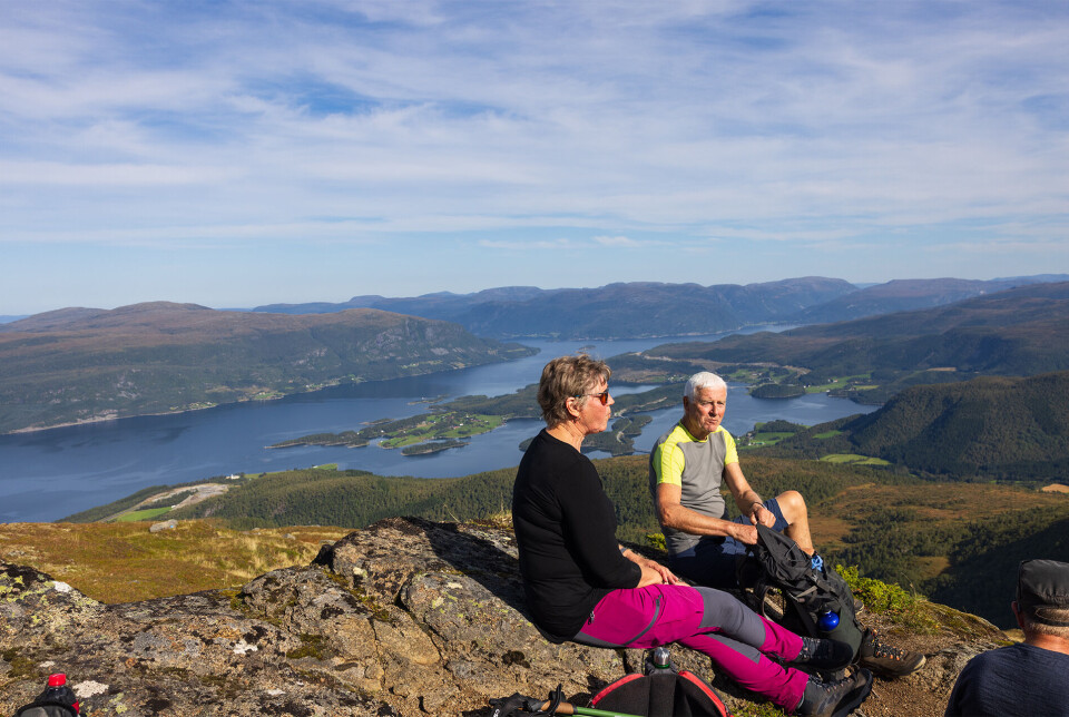 Matpause ved toppvarden. To turkledde personer, ei dame og en mann, som sitter ved siden av hverandre på en fjellknaus, med utsikt over fjord og fjell. Sommer og lettskyet himmel.