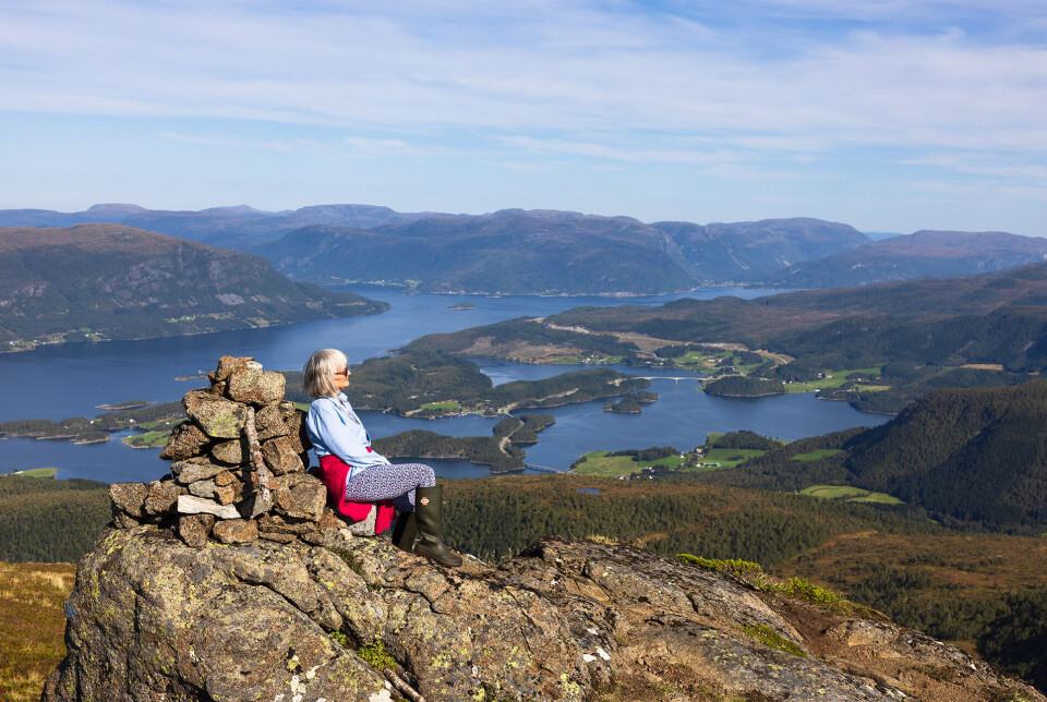 Flott utsyn over fjordar, øyer og fjell. Ei dame med halvlangt grått hår og solbriller, som sitter med ryggen lent mot en fjellvarde, med utsikt over fjord og fjell. Sommer og lettskyet himmel.