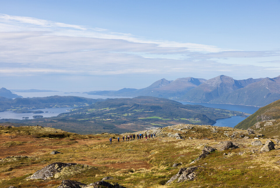 Fin nedtur med vidt og vakkert utsyn. Mange turkledde personer som går på rekke og rad etter hverandre på fjellet, med utsikt over fjord og fjell i bakgrunnen. Sommer og lettskyet himmel.