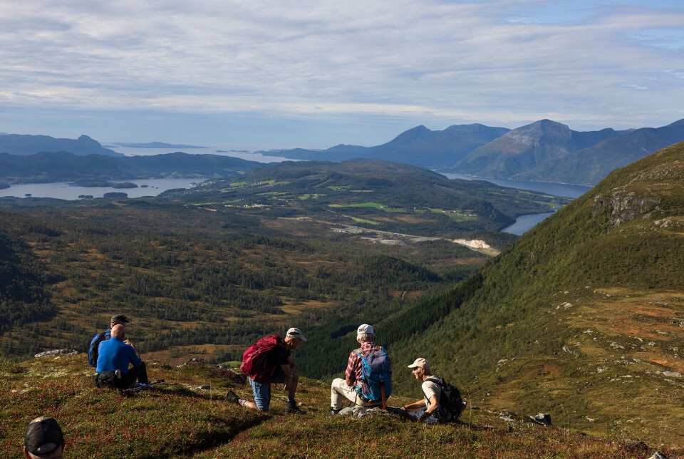 Liten kvil før den bratte lia ned. To turkledde personer, ei dame og en mann, som sitter ved siden av hverandre på en fjellknaus, med utsikt over fjord og fjell. Sommer og lettskyet himmel.