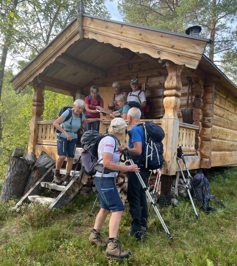 Gruvstuggu. Ei gruppe med turkledde folk ved ei tømmerhytte. Sommer, sol og lettskyet himmel.