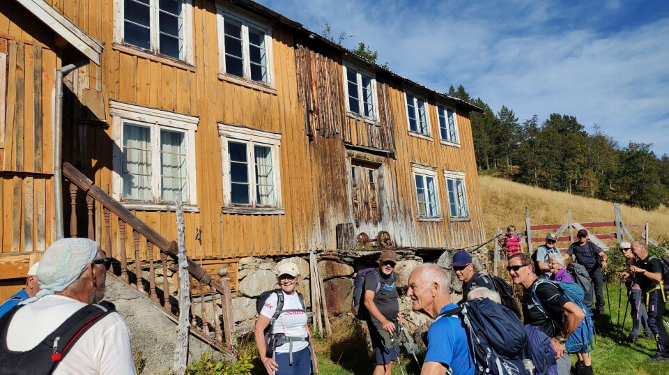 Høydalen. Ei gruppe med turkledde folk ved et gammelt gårdshus. Sommer, sol og lettskyet himmel.