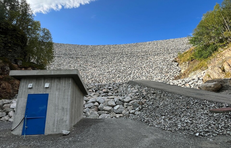 Lekkasjemålehus nedstrøms Follsjødammen. Nedsida av en stor demning på fjellet, dekket med store steiner. Sommer og blå himmel.